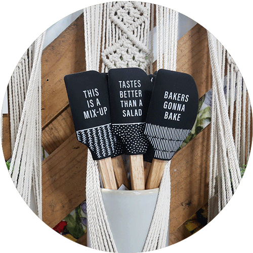 Three spatulas with sayings in a pot in a macrame holder. They say This Is A Mix-up, Tastes Better than a Salad, and Bakers Gonna Bake.